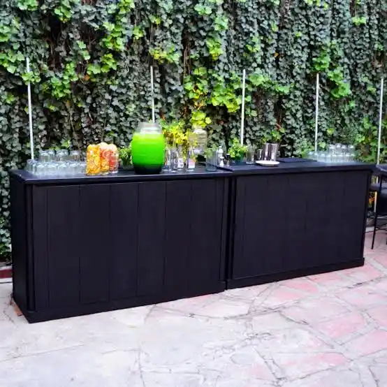 Barra de bebidas móvil de color negro con jarras de cócteles, cristalería y utensilios, instalada al aire libre frente a una pared cubierta de hiedra, ideal para eventos y fiestas.